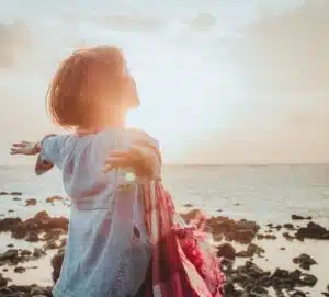 Woman standing on rocky beach wearing red scarf with arms outreached and sun brightly shining