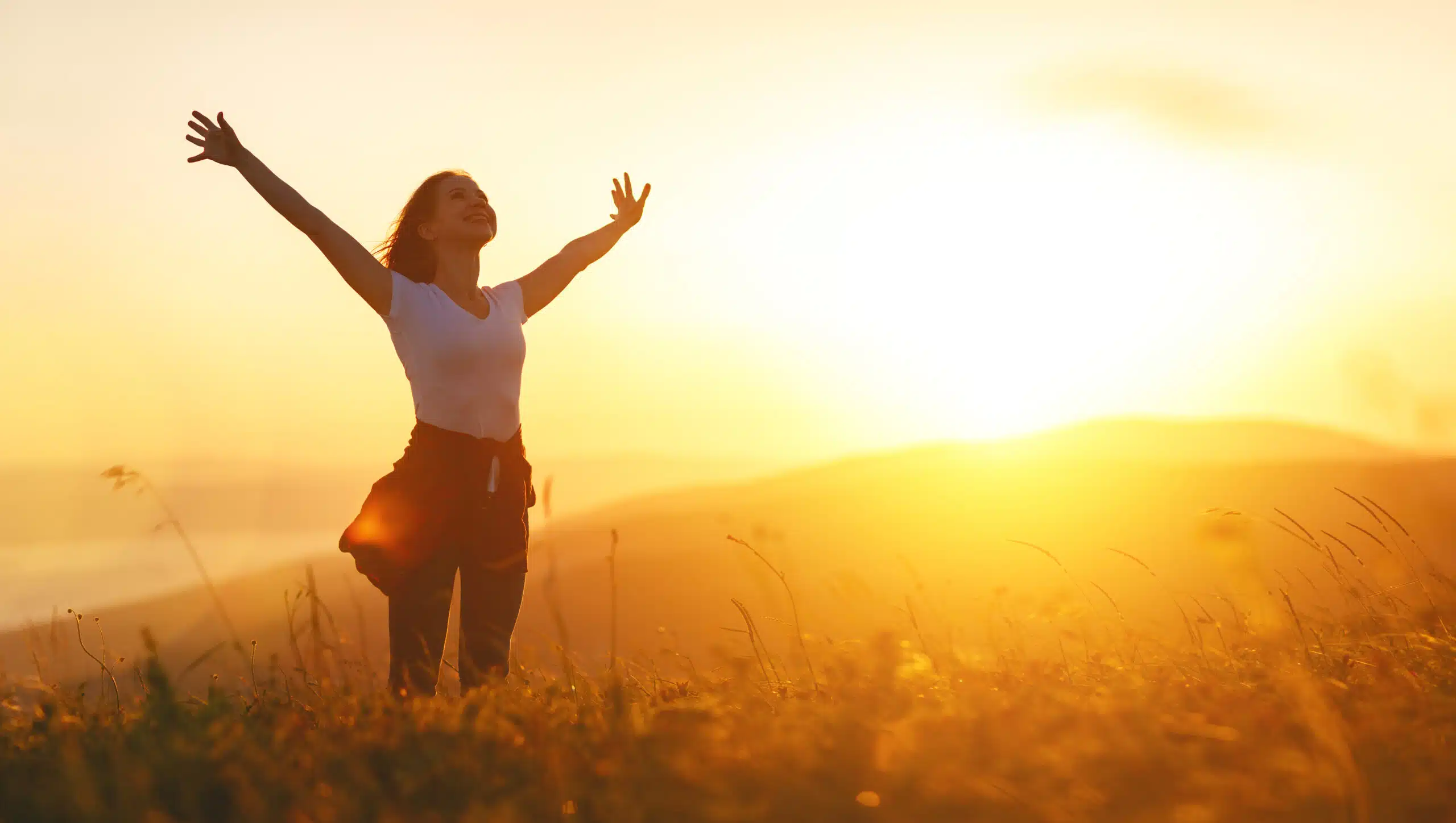 Woman smiling with arms outreached in a golden field with bright yellow sun shining in the background
