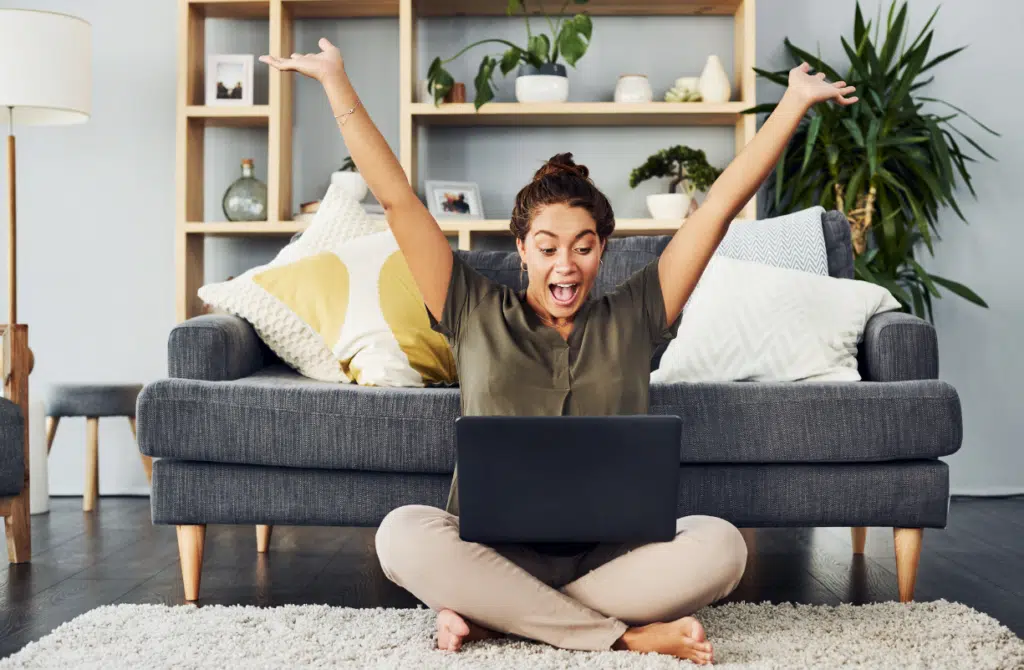 Woman of color sitting with legs crossed and hands up in the air as she looks with excitement at her laptop after learning these 7 Daily Habits for Success