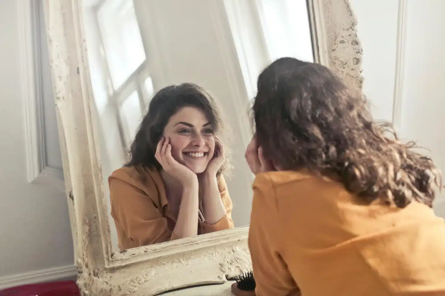 A woman in a warm-toned shirt smiles at her reflection in an antique mirror, embodying self-compassion with a gentle touch to her face, symbolizing positive self-regard and the practice of self-compassion.