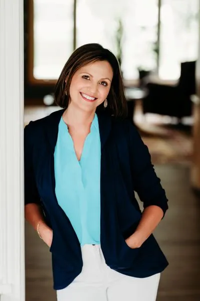 Dr. Stephanie Grunewald of Ancorio, smiling warmly while leaning against a doorway, wearing a turquoise blouse, navy blazer, and white pants.