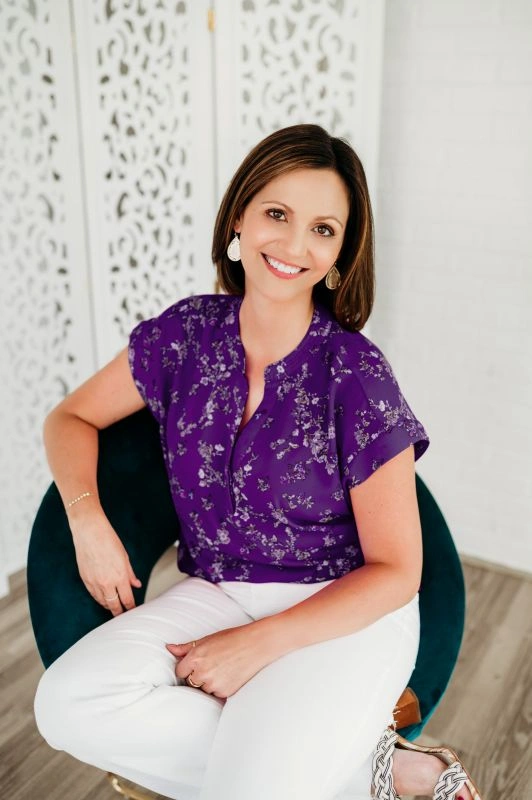 Dr. Stephanie Grunewald, Mindset & Alignment Strategist at Ancorio, seated in a teal chair wearing a purple floral shirt, smiling warmly.