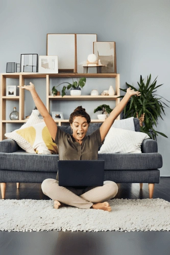 A woman sitting cross-legged on the floor in front of her laptop, excited and celebrating a personal win, representing the success and breakthroughs possible in the Total Life Reset program by Ancorio.