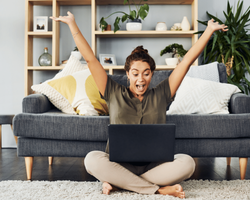 Woman of color sitting with legs crossed and hands up in the air as she looks with excitement at her laptop after learning these 7 Daily Habits for Success