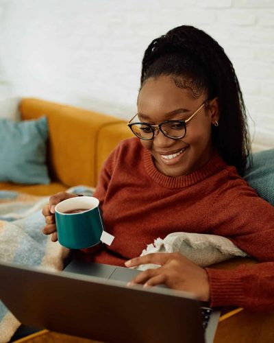 Happy African American woman drinking tea and using laptop while relaxing on the sofa.