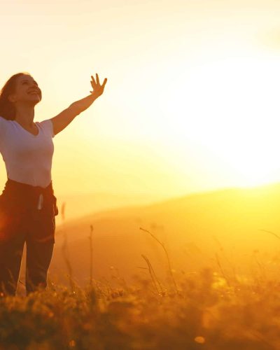 Woman smiling with arms outreached in a golden field with bright yellow sun shining in the background